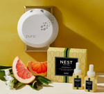 A white Pura smart home fragrance diffuser plugged into a gold-framed outlet on a vibrant yellow wall next to a Nest New York Grapefruit smart home fragrance diffuser refill box, two yellow individual refill bottles, fresh red grapefruit slices, and delicate white flowers with green leaves