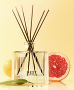 A NEST NEW YORK reed diffuser with black reeds in a clear rectangular bottle, flanked by a vibrant red grapefruit half and a yellow citrus fruit with a green leaf, all on a reflective surface against a soft yellow background