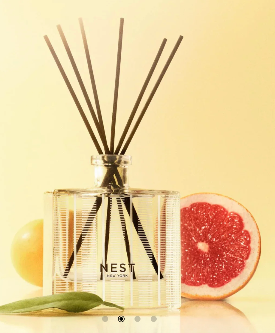 A NEST NEW YORK reed diffuser with black reeds in a clear rectangular bottle, flanked by a vibrant red grapefruit half and a yellow citrus fruit with a green leaf, all on a reflective surface against a soft yellow background