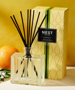 Product photograph, NEST New York, showcasing a Grapefruit Reed Diffuser in a striped glass bottle with black reeds, its yellow box featuring green vertical stripes and a subtle teardrop pattern, accompanied by a fresh grapefruit and white flowers against a vibrant yellow background