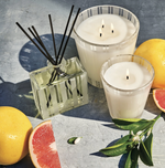 Product photography of a citrus-themed home fragrance display featuring a striped glass reed diffuser with black reeds, two lit striped glass candles (one triple-wicked, one single-wicked), a whole lemon, sliced pink grapefruit, and green citrus leaves with white blossoms on a textured blue surface