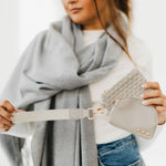 A woman wearing a light grey scarf and blue jeans holding a light grey canvas wristlet strap attached to two matching pouches, one with a woven texture and the other smooth leather embellished with three gold studs and bright gold hardware