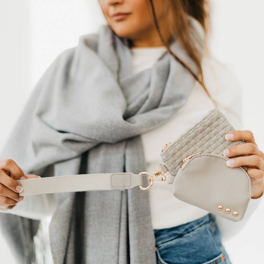 A woman wearing a light grey scarf and blue jeans holding a light grey canvas wristlet strap attached to two matching pouches, one with a woven texture and the other smooth leather embellished with three gold studs and bright gold hardware