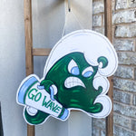 Painted cutout by A. DREZ of an angry green wave mascot with white foamy crests and blue eyes holding a white and blue "GO WAVE" megaphone hanging on a wooden ladder outdoors against stucco and brick walls