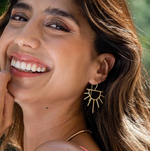 Close-up of a smiling woman with warm brown hair and shimmering eye makeup wearing gold sunburst earrings in natural outdoor light