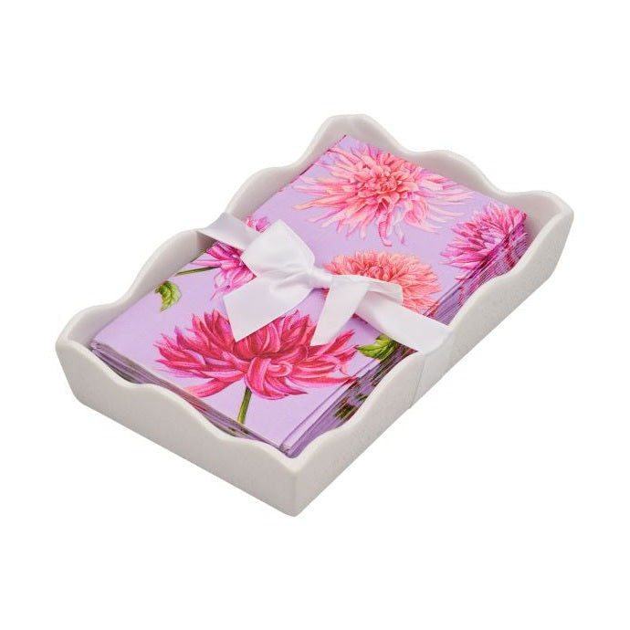 A stack of purple napkins with vibrant pink dahlia illustrations, tied with a white ribbon, sitting in a white wavy ceramic napkin holder