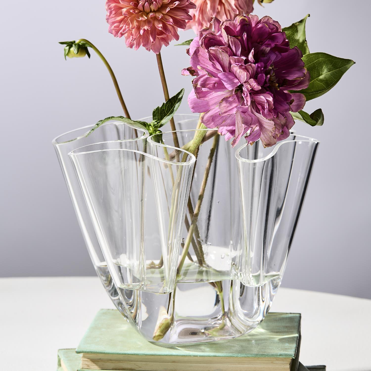 A clear wavy glass vase holds vibrant pink and faded purple dahlias and a bud, resting on a stack of light green vintage books on a white table