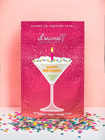Product photograph by d'marie featuring a Happy Birthday cocktail slush mix pouch on a pink backdrop with a martini glass graphic, a lit pink candle, and multicolored sprinkles above scattered confetti