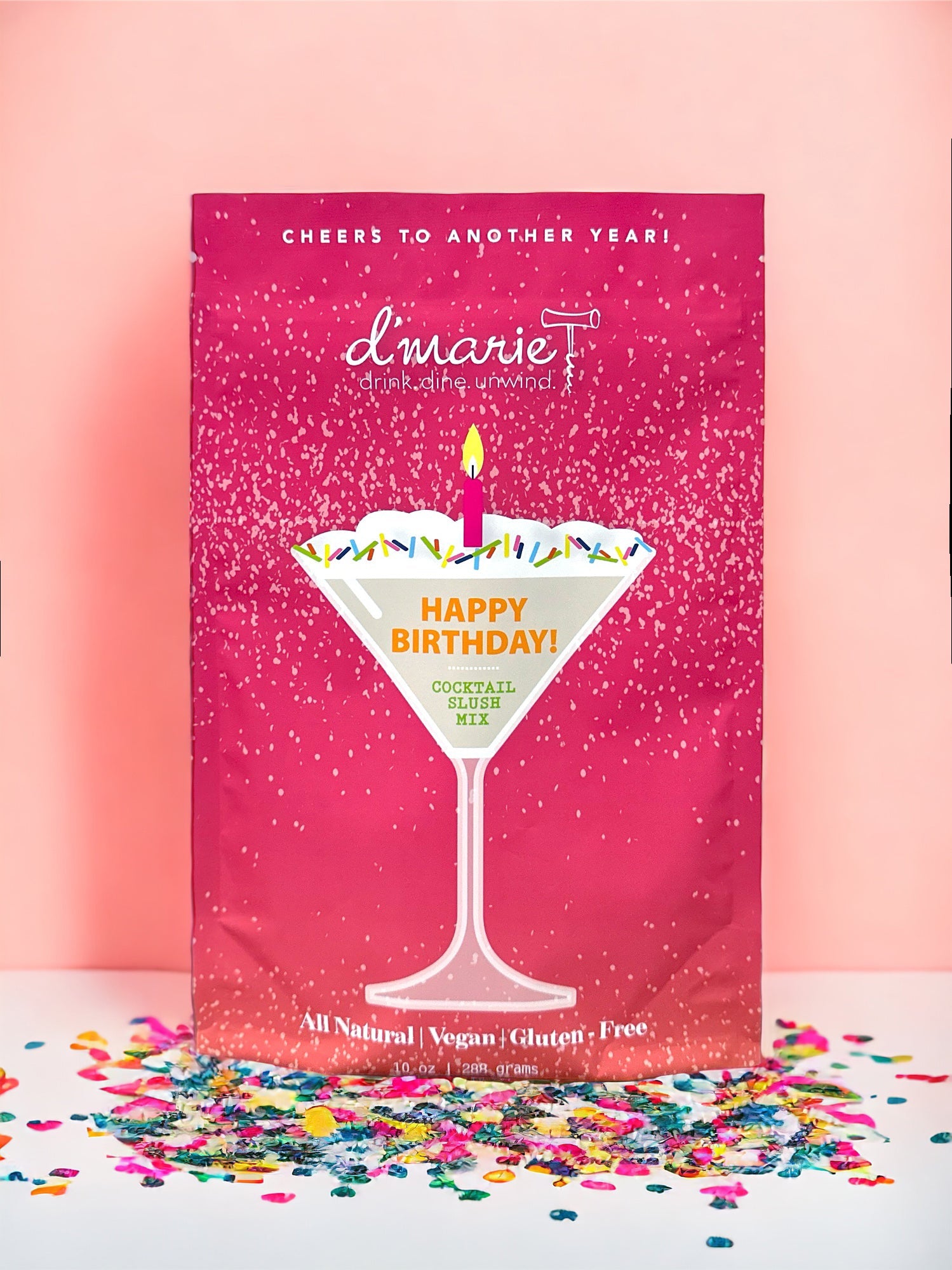 Product photograph by d'marie featuring a Happy Birthday cocktail slush mix pouch on a pink backdrop with a martini glass graphic, a lit pink candle, and multicolored sprinkles above scattered confetti