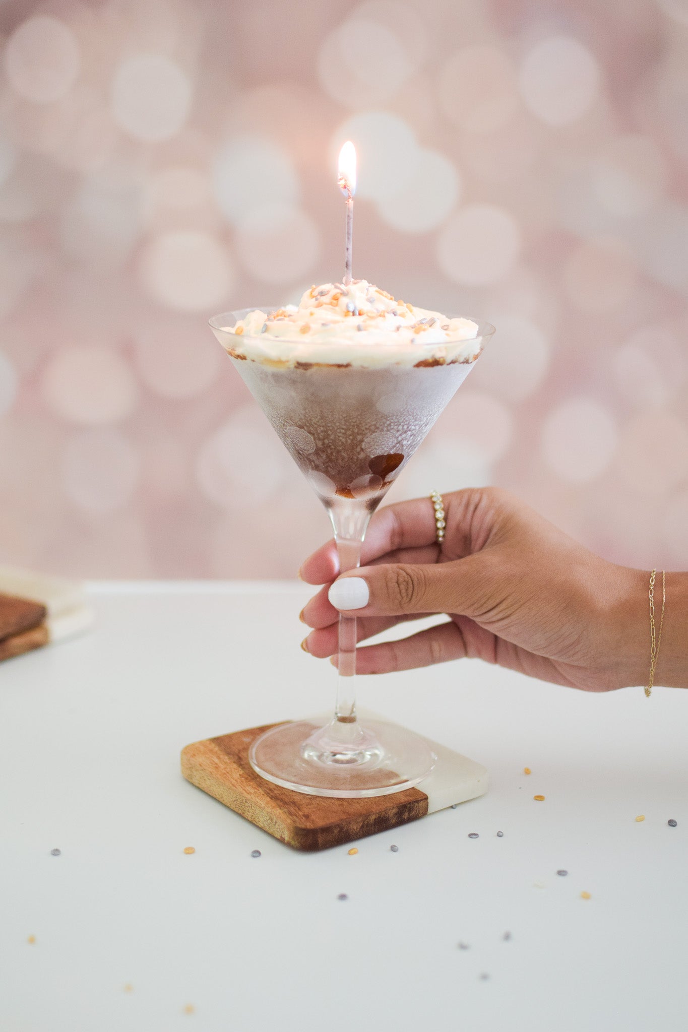 Photograph of a manicured hand holding a frosted martini glass topped with whipped cream and a lit candle on a wood and marble coaster against a soft pink bokeh background