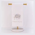 Embroidered white tea towel featuring a sketch of a New Orleans French Quarter building with iconic cast-iron balconies draped with colorful purple green and yellow Mardi Gras beads and "happy mardi gras" text elegantly displayed on a brass and marble stand