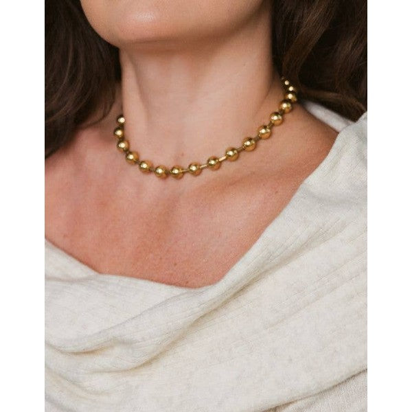 Photograph of a woman showcasing a gold-tone spherical bead choker necklace against a cream ribbed sweater with a draped neckline