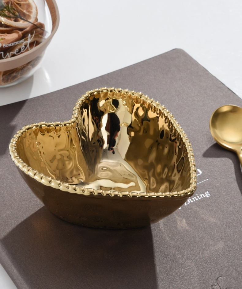 A shiny hammered gold heart-shaped bowl with a beaded rim rests on a dark placemat alongside a matching gold spoon and a clear bowl of dried citrus slices