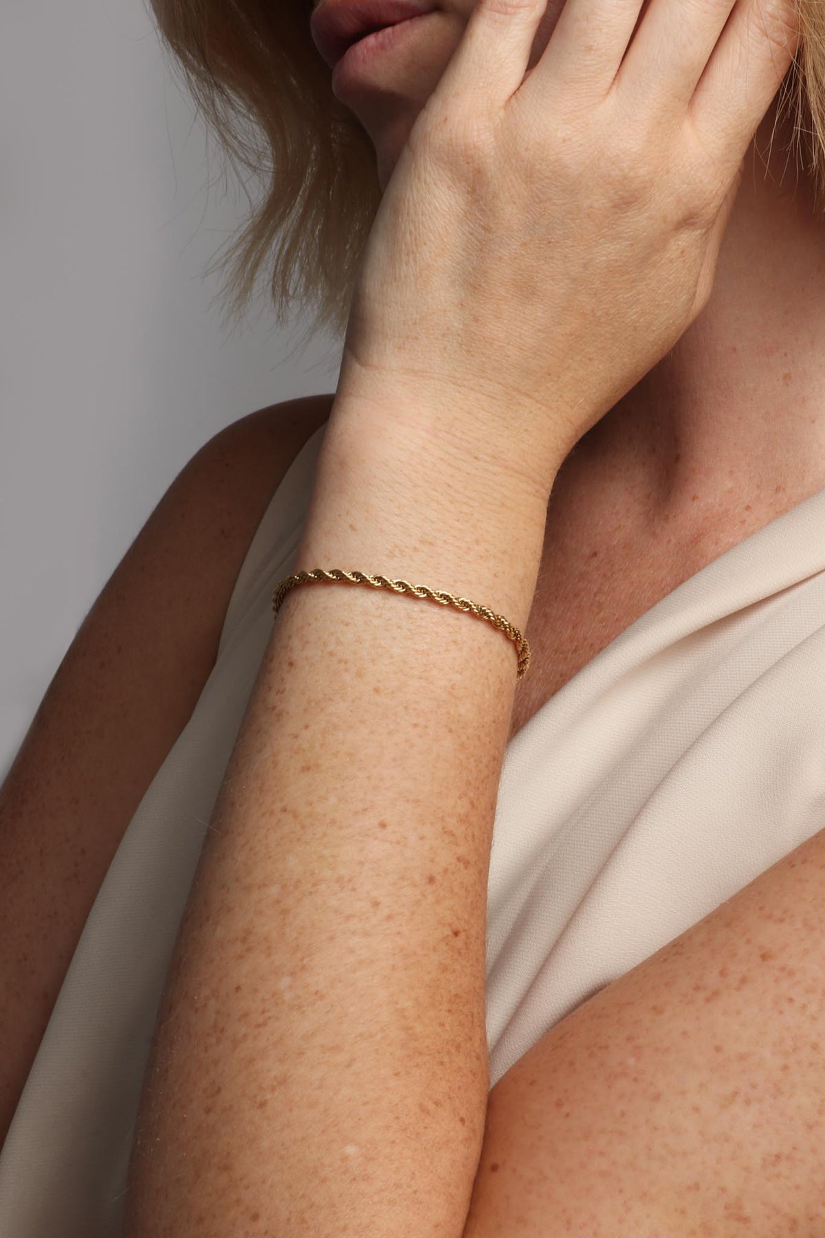 Close-up of a woman's freckled arm adorned with a golden rope chain bracelet, her blonde hair and hand partially obscuring her face against a cream-colored fabric