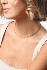 A woman's décolletage with short blonde hair and freckled skin wearing two delicate gold layered necklaces, one a twisted rope chain and the other a small beaded satellite chain, against a light beige tank top