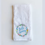 White waffle weave towel embroidered with "Hello Spring" in blue script encircled by a colorful wreath of pink, yellow, and blue flowers