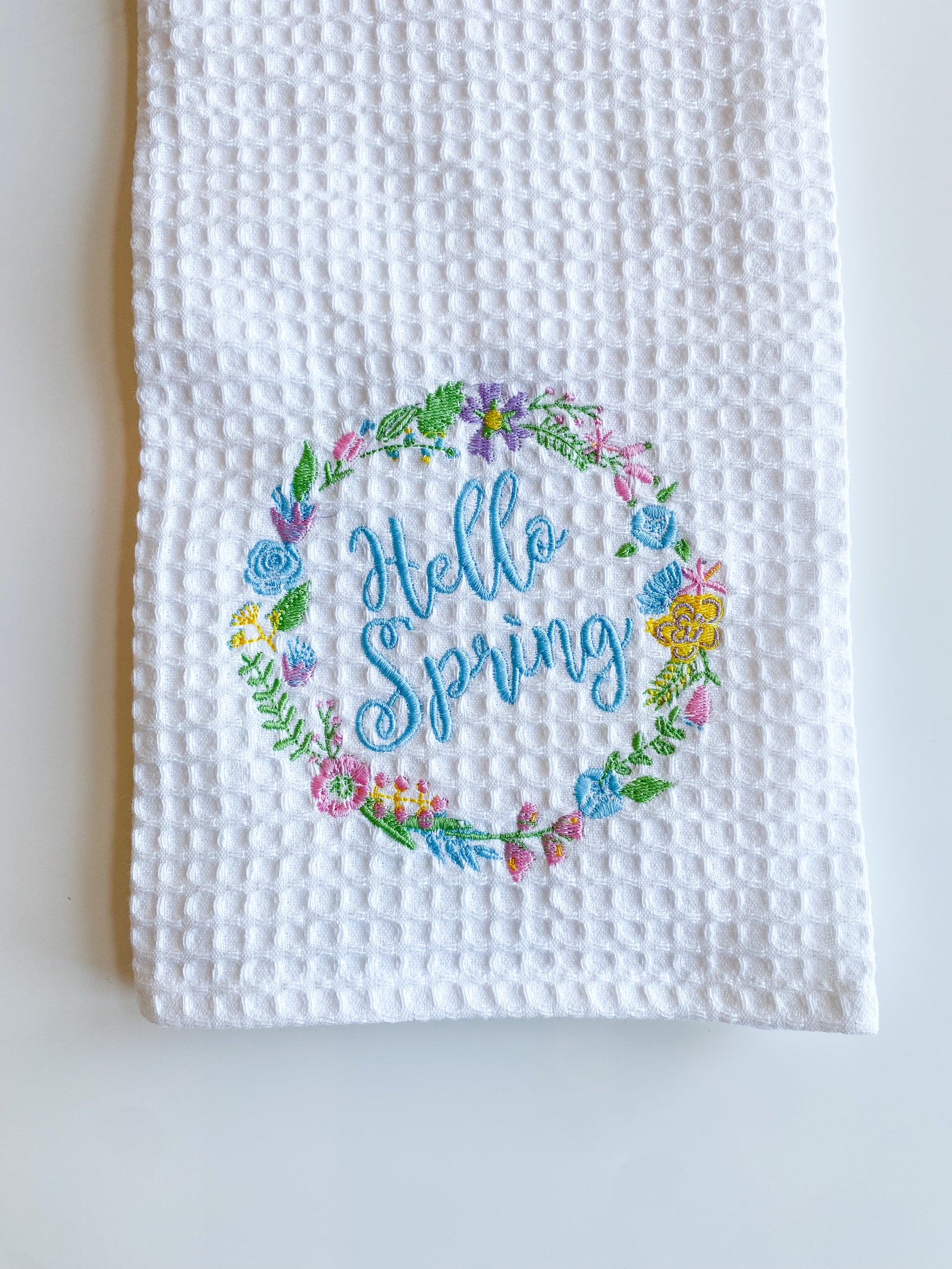 Embroidered white waffle weave towel featuring a vibrant floral wreath with pink blue yellow and purple flowers framing light blue Hello Spring script