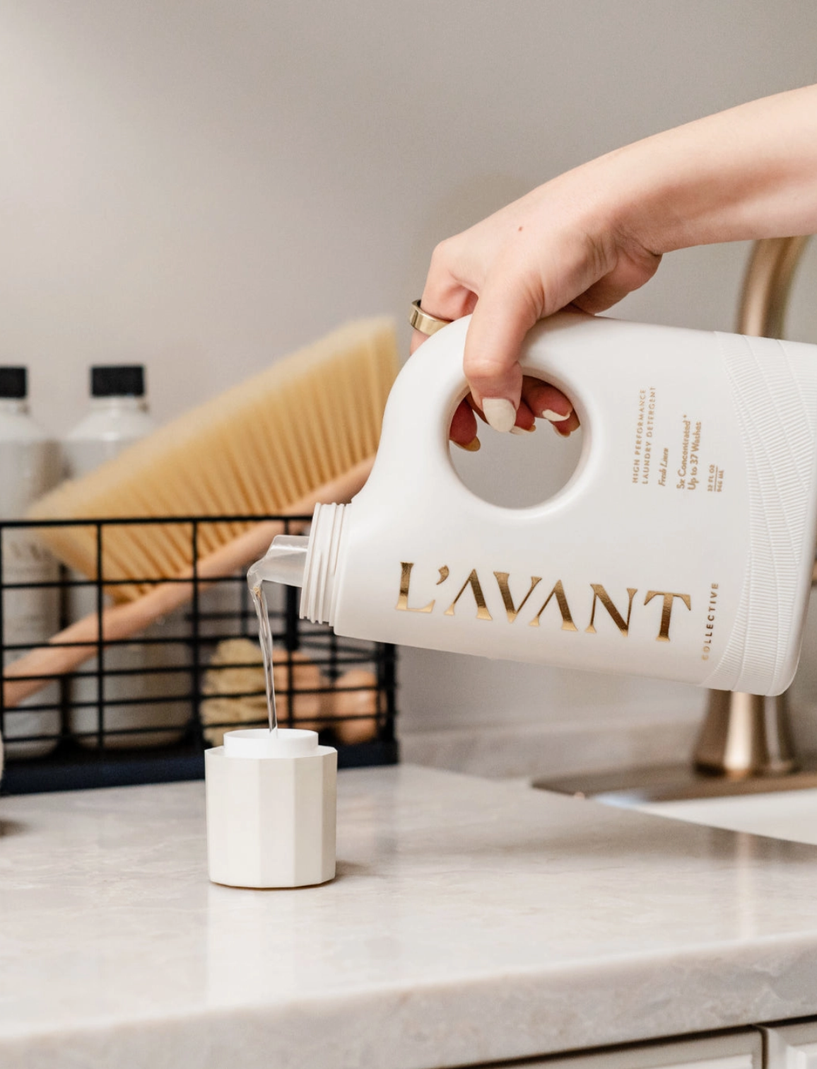 A hand pours clear L'AVANT Collective laundry detergent from a white bottle with gold lettering into a faceted white measuring cap on a white countertop, featuring a soft-focus gold faucet and wooden brush in the background