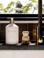 Photograph of a L'AVANT Collective white dish soap bottle with a gold pump and a natural wood brush on a dark kitchen counter with a gold faucet and green leaves visible outside