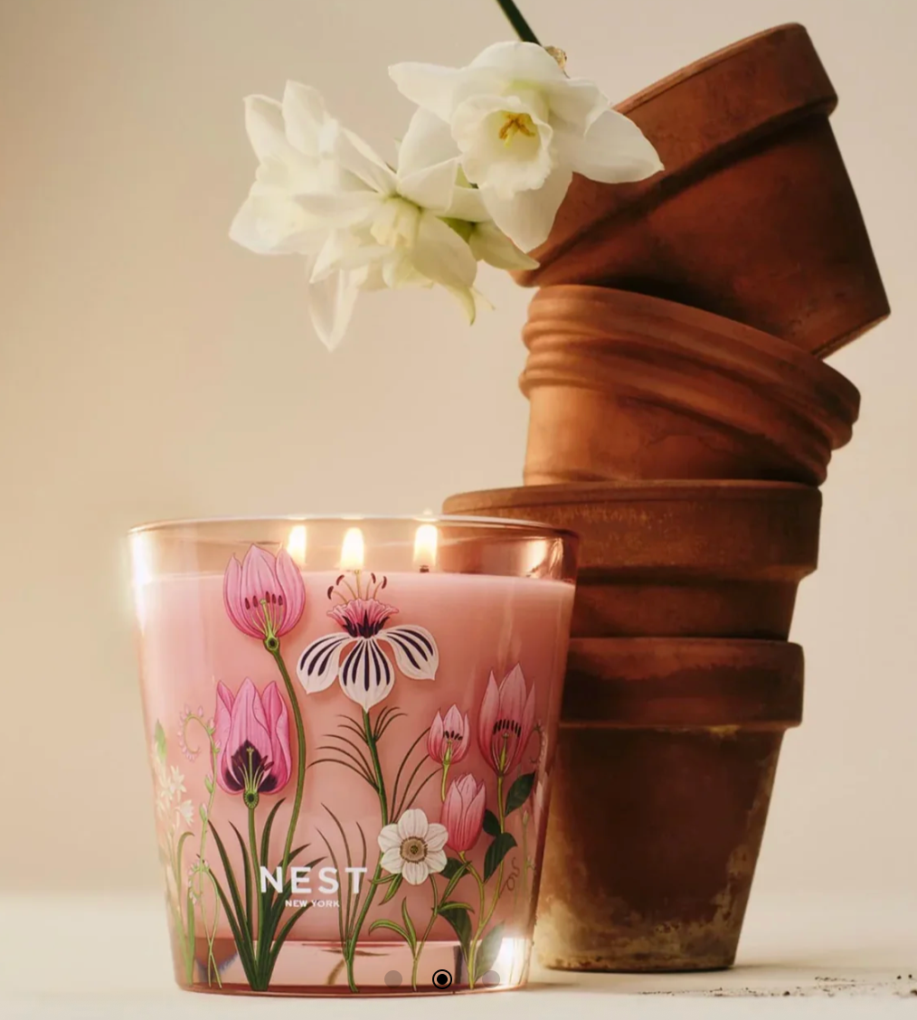 Photograph by NEST NEW YORK featuring a glowing three-wick pink candle in a glass jar with vibrant floral illustrations of pink tulips and a striped bloom, next to stacked terracotta pots topped with delicate white daffodils