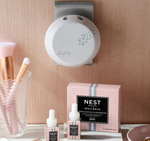 Photograph of a modern white Pura smart home diffuser mounted on a rose gold textured wall, alongside Nest New York Wellness Himalayan Salt & Rosewater fragrance refills and blush pink makeup brushes in a clear glass on a white marble surface