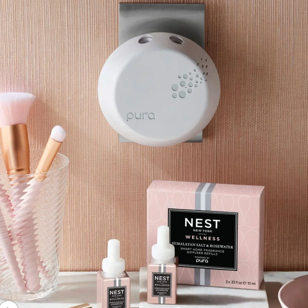Commercial photograph for Pura and Nest New York featuring a white circular smart fragrance diffuser with bubble-patterned vents plugged into a tan ribbed wall above a pink Nest Wellness box and soft pink makeup brushes on a white marble vanity