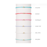 Six colorful beaded bracelets with gold beads displayed on a white stand with color labels
