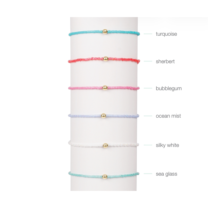 Six colorful beaded bracelets with gold beads displayed on a white stand with color labels