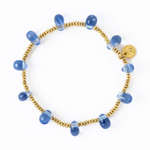 A bracelet featuring small gold-colored round beads interspersed with larger teardrop-shaped translucent blue glass beads each with a white stripe and a small round gold-toned charm with an engraved design