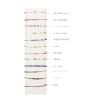 A collection of colorful seed bead bracelets displayed vertically on a white stand each featuring a central gold bead and varied designs including rainbow multi-color, vibrant fuchsia, and deep blue metallic styles
