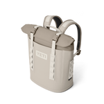 Digital render of a tan YETI Hopper M20 backpack soft cooler featuring a prominent YETI logo, roll-top closure with white straps and buckles, and padded shoulder straps against a black studio background