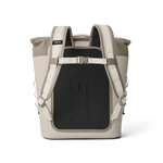 Yeti product photo of a two-tone tan and taupe roll-top backpack cooler on a black background, featuring white padded shoulder straps, a chest buckle, and an embossed Yeti logo on the black back panel