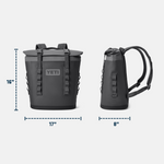 Studio product photo of a two-tone dark and light grey YETI Hopper M12 backpack cooler on a plain background, featuring a rolltop closure, prominent YETI logo, and dimensions 16 inches high 17 inches wide and 8 inches deep