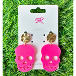 Acrylic and metal earrings by SR (Sandy Rizzolo) featuring neon pink skull cutouts with black eye sockets and teeth, dangling from round textured gold stud tops, presented on a white display card with a pink SR logo against an artificial green grass background