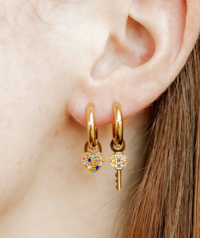 A close-up of an ear wearing gold hoop earrings with a dangling heart-shaped padlock charm and a key charm, both intricately studded with colorful sparkling gemstones