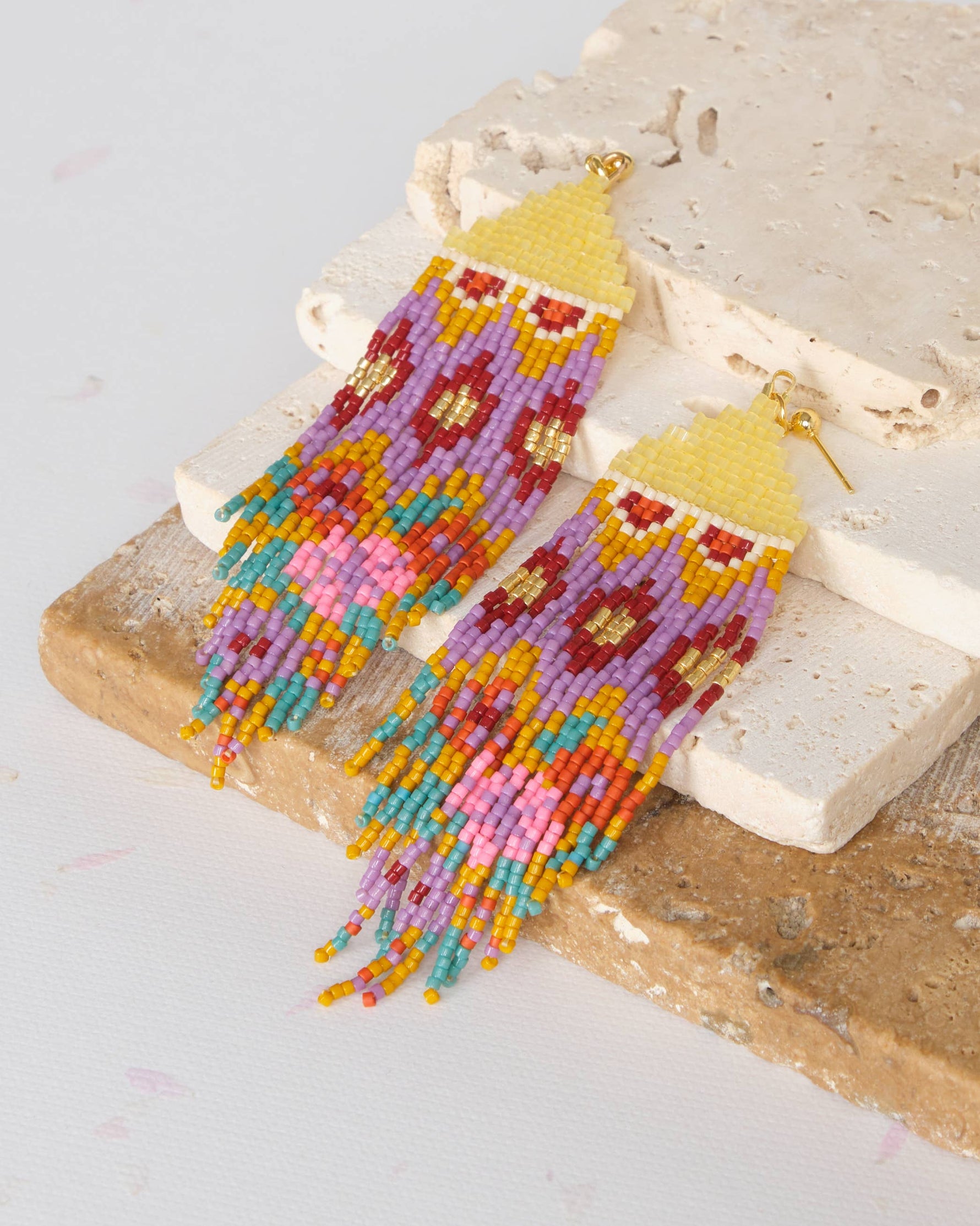 Handcrafted beaded fringe earrings in a vibrant color palette of yellow, purple, gold, red, teal, orange, and pink with geometric patterns, resting on textured stone slabs