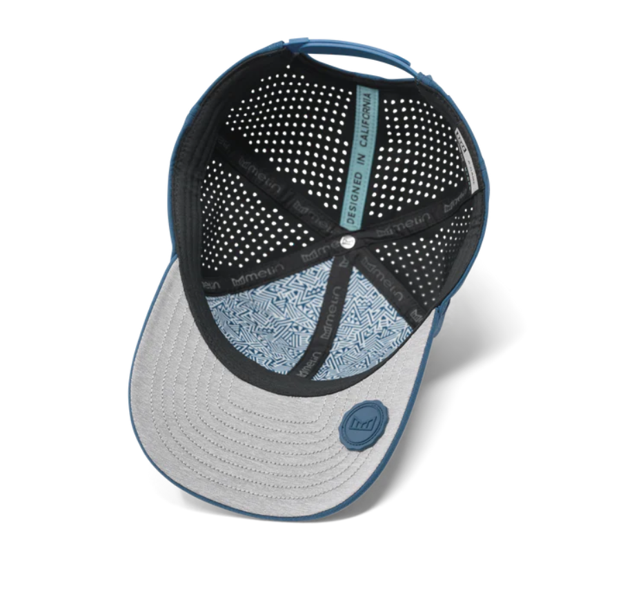 Melin blue performance baseball cap viewed from the underside, revealing a perforated black mesh interior, a grey brim with concentric stitching, blue geometric patterned lining, and a blue rubber Melin logo badge with a "DESIGNED IN CALIFORNIA" tag