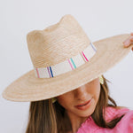 A woman in a bright pink top wearing a natural straw fedora hat with a cream band featuring colorful vertical stripes of blue, purple, pink, and green, her face partially obscured by the brim and subtle yellow citrus slice earrings visible
