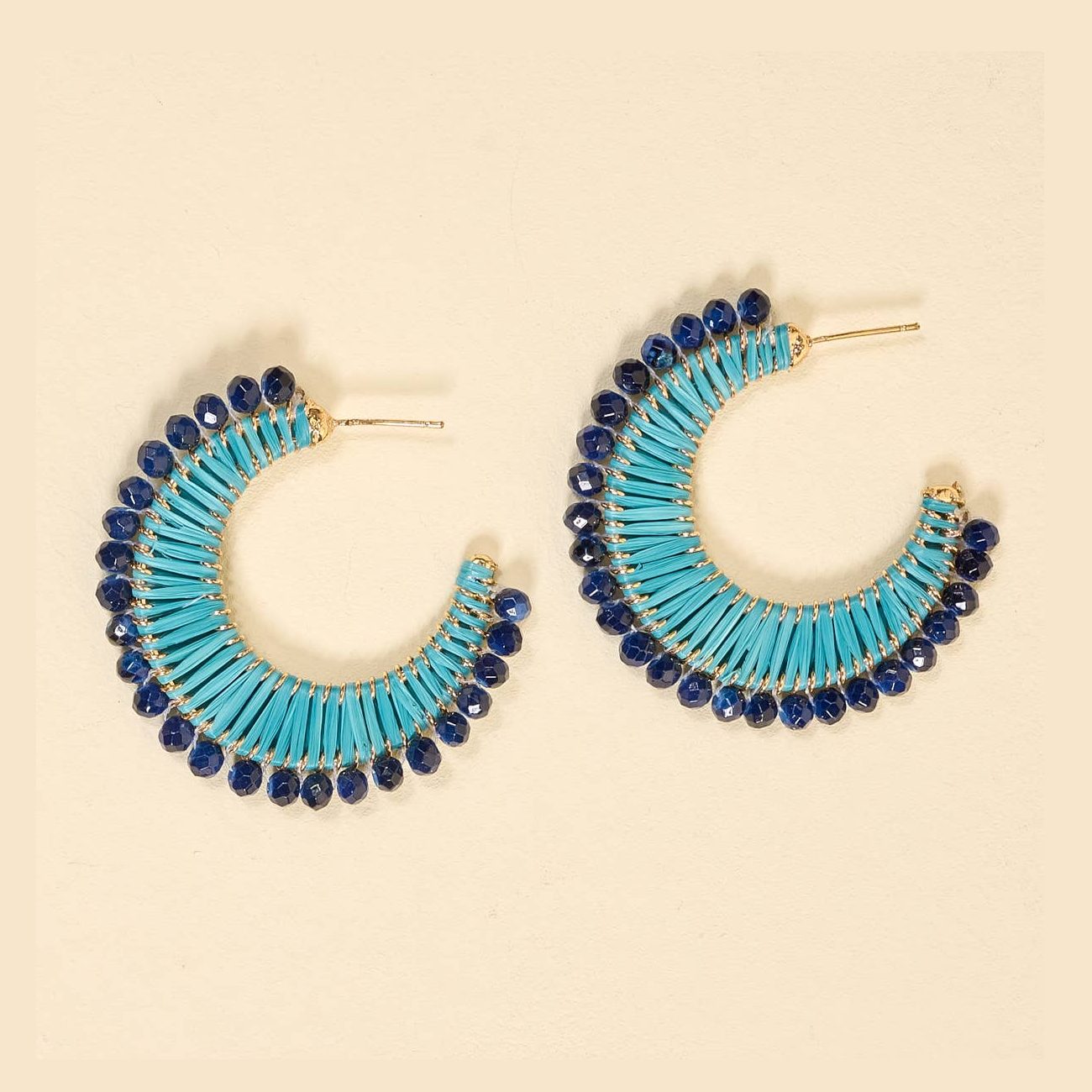 A pair of crescent-shaped hoop earrings featuring bright turquoise woven raffia and dark blue faceted bead embellishments
