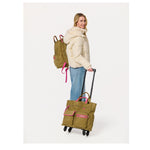 Woman wearing a cream puffer jacket and jeans carrying an olive green backpack and rolling an olive green suitcase