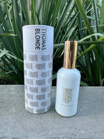Product photography of Thomas Blonde Parfum D'Interieur fragrance bottle and its white cylindrical box featuring 'THOMAS BLONDE' text, displayed on a grey concrete surface with lush green foliage in the background, both items prominently showcasing a gold TB monogram and the bottle topped with a reflective gold cap