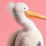 A white fluffy plush pelican toy with a long orange beak, a spiky tuft of white feathers, and a hint of dark grey details against a vibrant pink background