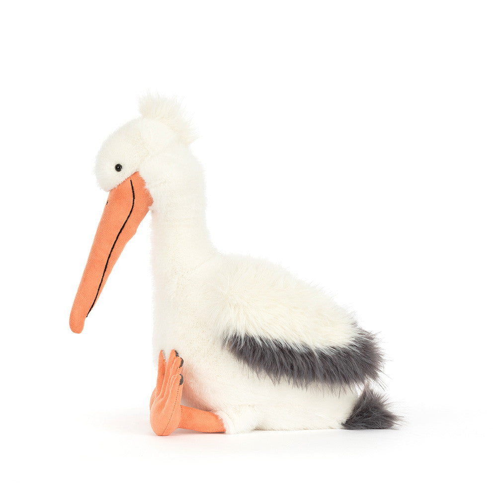 Plush toy by Jellycat of a sitting pelican on a white background featuring a long orange beak, charcoal grey wing tips, and a tufted white crest