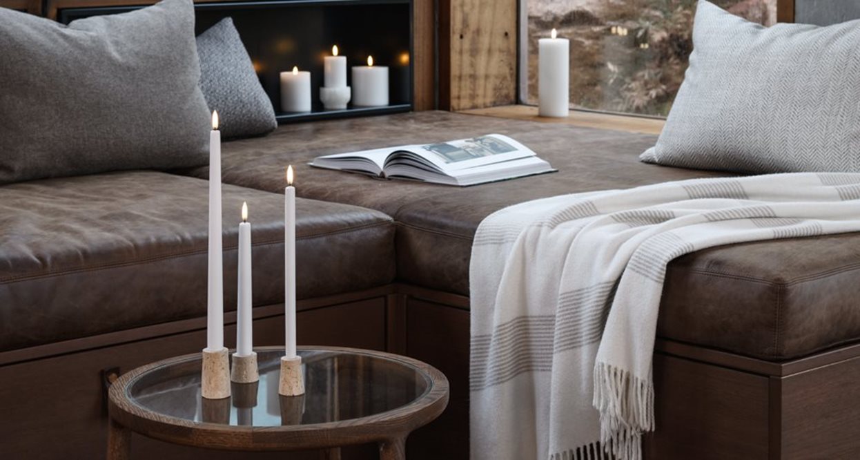 Three lit taper candles on a glass table next to a brown leather sofa with cushions and a throw blanket