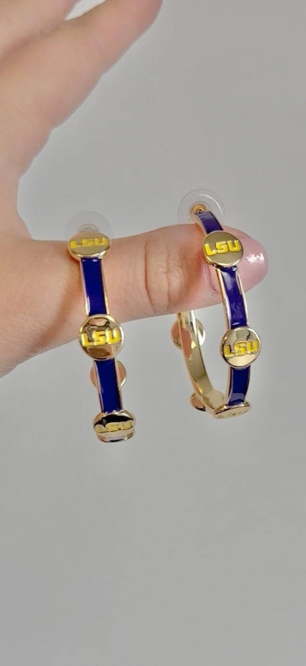 A pair of shiny gold hoop earrings featuring deep purple enamel bands and multiple yellow LSU logos held by a hand