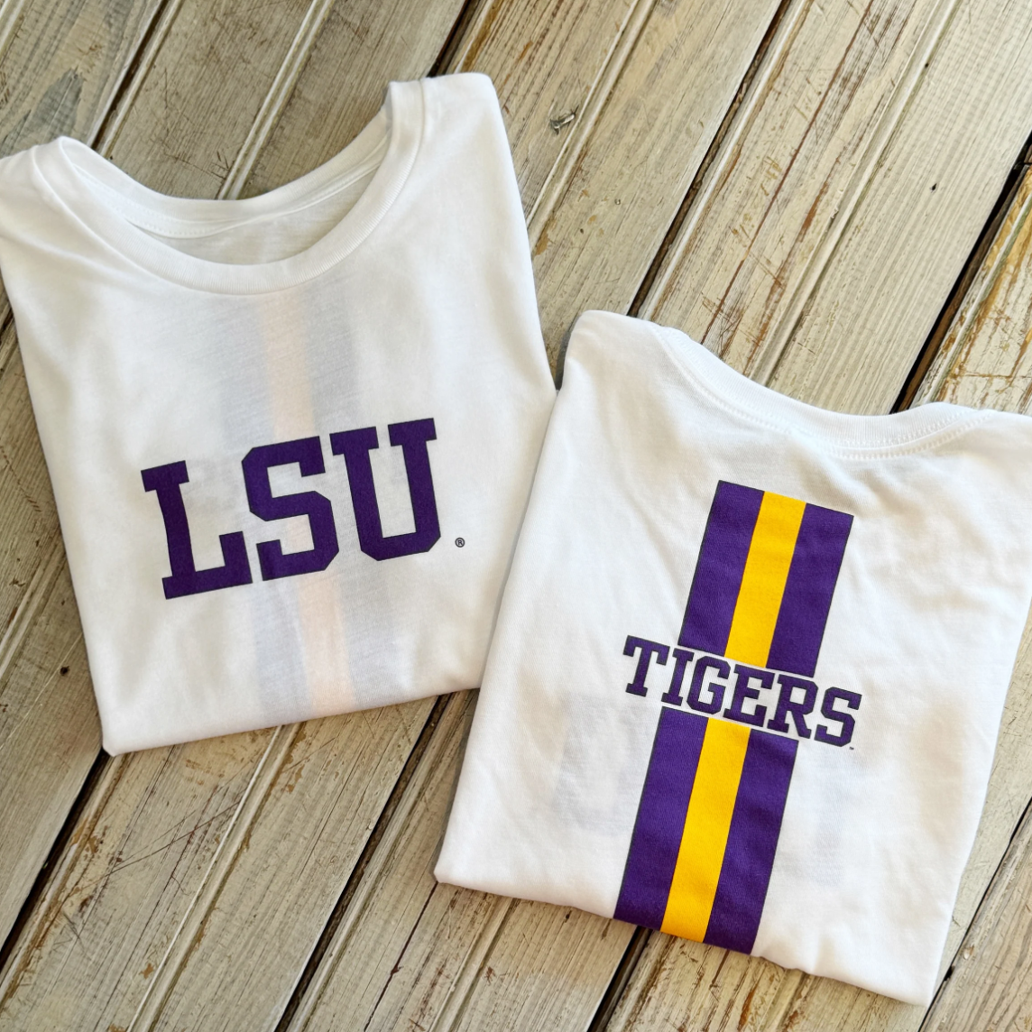 Two white t-shirts with purple and yellow LSU Tigers graphics on a wooden background