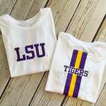 Two white LSU branded t-shirts one featuring bold purple LSU lettering and the other displaying TIGERS over a vibrant vertical purple and gold stripe laid on a weathered light wood surface