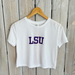 Photograph of a white crop top t-shirt featuring bold purple LSU text hanging on a wooden hanger against a distressed light gray vertical plank wood wall