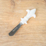 Stainless steel and white ceramic butter spreader featuring a decorative fleur-de-lis handle on a textured light wood surface with a rounded blade inscribed with STAINLESS STEEL MADE IN CHINA