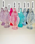 Eight translucent glass Virgin Mary statues in various colors labeled with names